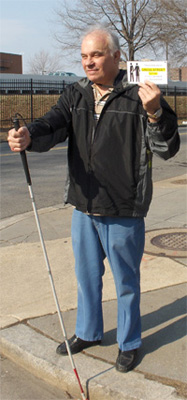 A deaf-blind man, Art, smiles and stands at the curb with his left hand holding the card above his left shoulder, to be seen from front and back.  His right arm is extended forward to hold a cane, the tip of the cane is at the curb.